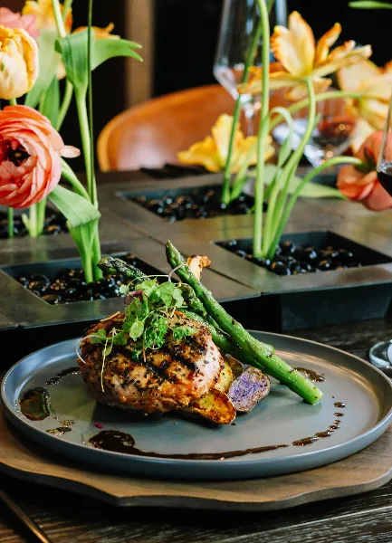 A plated meal consisting of a seared chicken breast, grilled asparagus, and microgreens served on a grey plate with a floral centerpiece in the background.