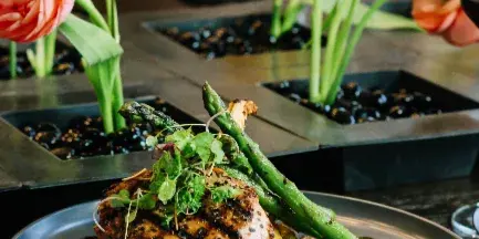 A plated meal consisting of a seared chicken breast, grilled asparagus, and microgreens served on a grey plate with a floral centerpiece in the background.