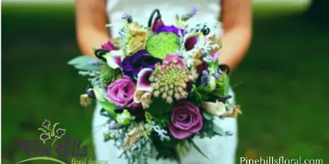 A bride holding a vibrant purple and pink wedding bouquet with the Pine Hills Floral Design logo in the corner.