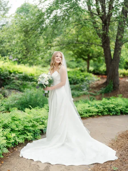 A bride in a white gown and veil holding a bouquet of white flowers, standing outdoors in a garden setting.