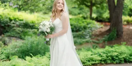 A bride in a white gown and veil holding a bouquet of white flowers, standing outdoors in a garden setting.