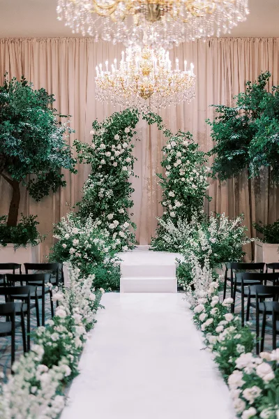 Indoor wedding ceremony setup with a floral arch, white aisle, chandeliers, and rows of chairs.