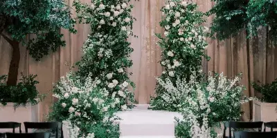 Indoor wedding ceremony setup with a floral arch, white aisle, chandeliers, and rows of chairs.