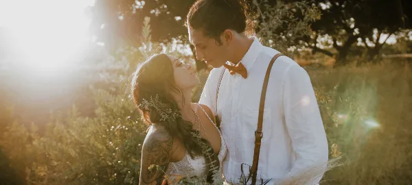 A bride and groom in wedding attire standing close together in a sunlit field with greenery in the background.