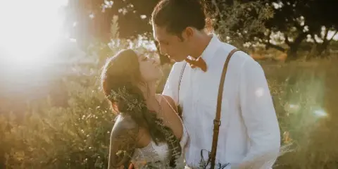 A bride and groom in wedding attire standing close together in a sunlit field with greenery in the background.