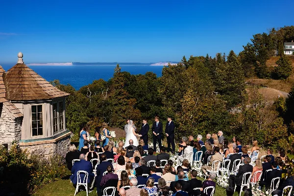 A wide shot of a wedding ceremony taking place outdoors on a hillside overlooking a large lake, with the bride and groom at the altar and guests seated in rows of white chairs.