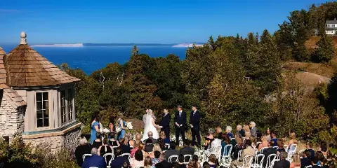 A wide shot of a wedding ceremony taking place outdoors on a hillside overlooking a large lake, with the bride and groom at the altar and guests seated in rows of white chairs.