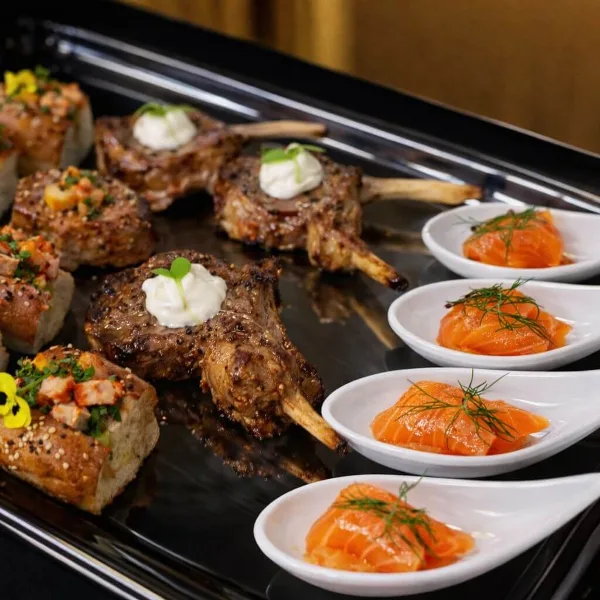 Catering platter featuring grilled lamb chops, smoked salmon portions, and bread appetizers on a black serving tray.
