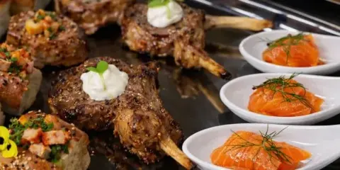 Catering platter featuring grilled lamb chops, smoked salmon portions, and bread appetizers on a black serving tray.