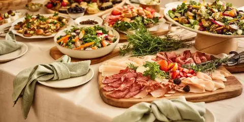 A catering table display featuring a wooden board with sliced cured meats, fresh vegetable salads in white bowls, and other appetizers.
