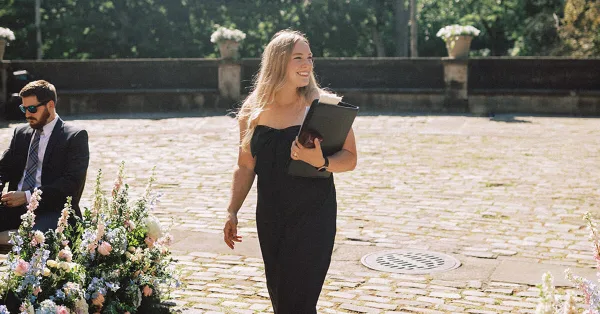 A woman in a black dress holding a binder at an outdoor wedding ceremony with floral arrangements.