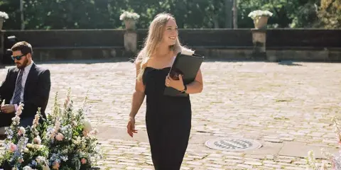 A woman in a black dress holding a binder at an outdoor wedding ceremony with floral arrangements.
