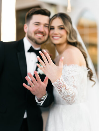 A bride and groom smiling at the camera, with the bride holding up her hand to show her wedding ring.