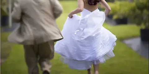 A bride in a white dress and a groom in a tan suit walking through a garden.