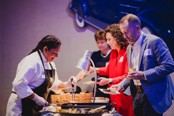 A chef in a white uniform and black apron serves food from a buffet station to guests at an event.