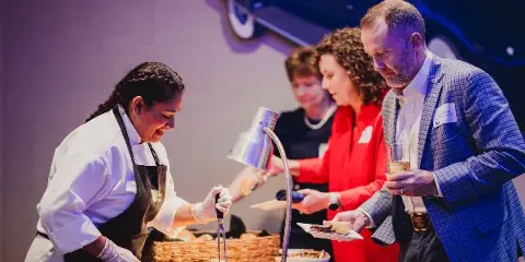 A chef in a white uniform and black apron serves food from a buffet station to guests at an event.