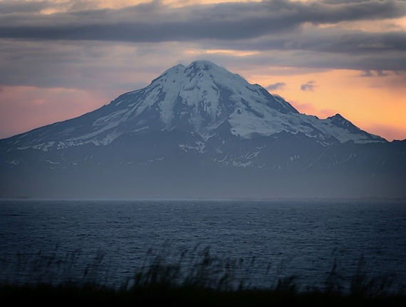 A snow-capped mountain viewed across a body of water at sunset with a grassy foreground.