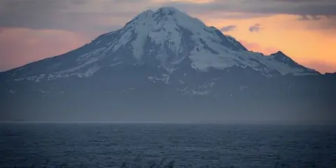 A snow-capped mountain viewed across a body of water at sunset with a grassy foreground.