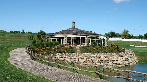 Exterior view of the Villa on the Green event venue featuring a stone chimney, large glass windows, and a wooden walkway leading to the entrance.