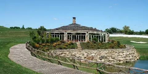 Exterior view of the Villa on the Green event venue featuring a stone chimney, large glass windows, and a wooden walkway leading to the entrance.