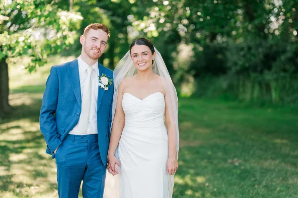 A bride in a white gown and a groom in a blue suit standing together outdoors.
