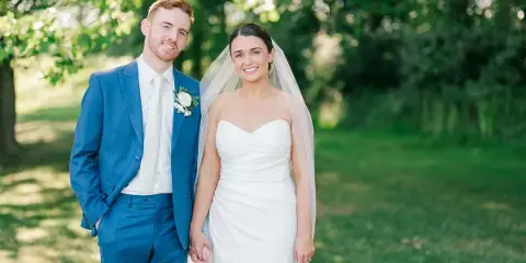 A bride in a white gown and a groom in a blue suit standing together outdoors.