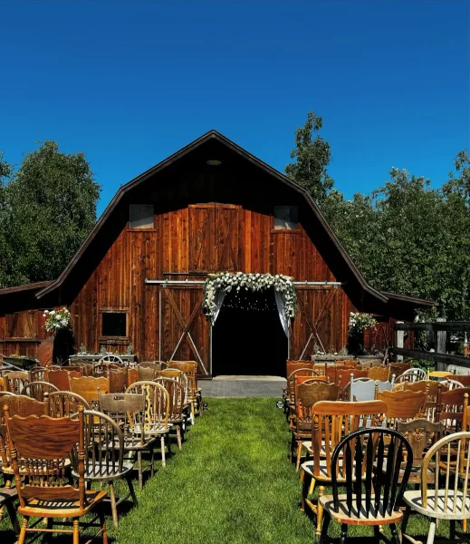 A wedding ceremony setup with rows of wooden chairs and a floral arch in front of a rustic wooden barn.