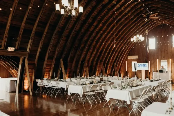 Interior view of a rustic wooden event venue with arched ceilings, chandeliers, and set reception tables.