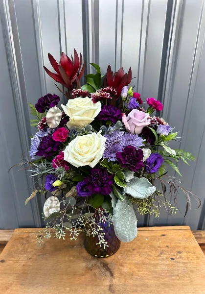 A professional floral arrangement featuring white and purple roses, carnations, and eucalyptus in a glass vase on a wooden table.
