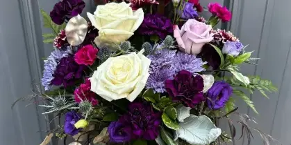 A professional floral arrangement featuring white and purple roses, carnations, and eucalyptus in a glass vase on a wooden table.