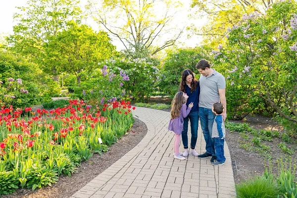 A family of four posing on a brick path in a garden with blooming red tulips and lilac bushes.