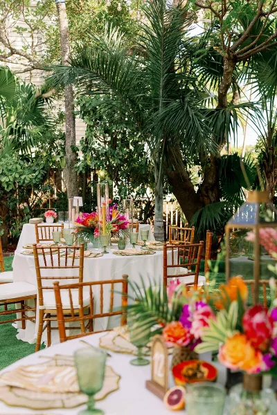 A wedding reception table set with white linens, bamboo chairs, and vibrant tropical floral arrangements in a garden setting.