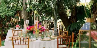 A wedding reception table set with white linens, bamboo chairs, and vibrant tropical floral arrangements in a garden setting.