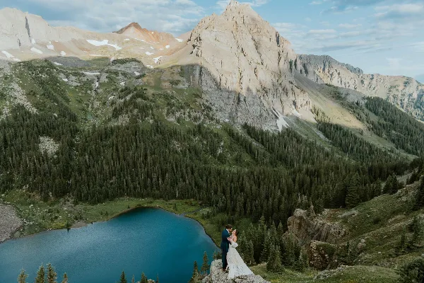 A bride and groom standing on a cliff edge overlooking a turquoise mountain lake and rugged peaks.