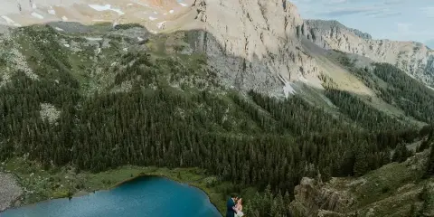 A bride and groom standing on a cliff edge overlooking a turquoise mountain lake and rugged peaks.