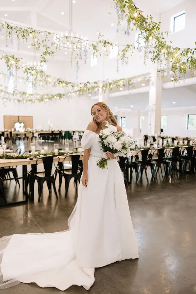 A bride in a white gown holding a bouquet stands in a spacious, modern barn-style wedding venue with long tables and hanging floral decor.