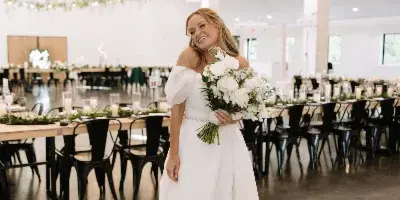 A bride in a white gown holding a bouquet stands in a spacious, modern barn-style wedding venue with long tables and hanging floral decor.