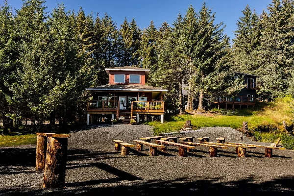 A two-story wooden cottage with a deck and outdoor picnic tables situated in a forested area.
