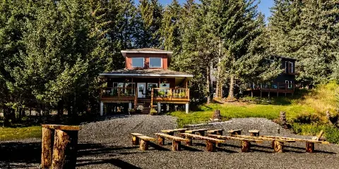 A two-story wooden cottage with a deck and outdoor picnic tables situated in a forested area.