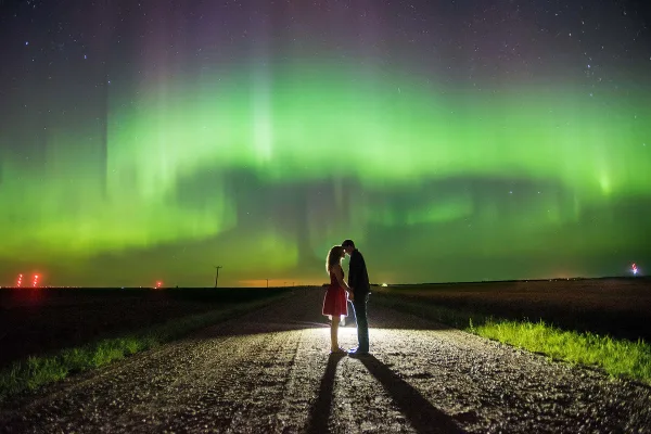 A couple standing on a rural road at night with the green aurora borealis visible in the sky.