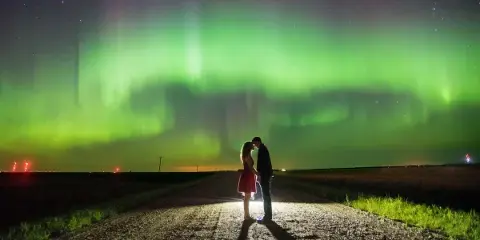 A couple standing on a rural road at night with the green aurora borealis visible in the sky.