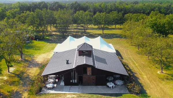 Aerial view of a large barn-style event venue with a white tent and set-up tables on a grassy estate.