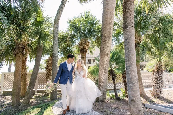 A bride and groom standing together outdoors among palm trees, with the bride holding a bouquet.