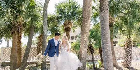 A bride and groom standing together outdoors among palm trees, with the bride holding a bouquet.