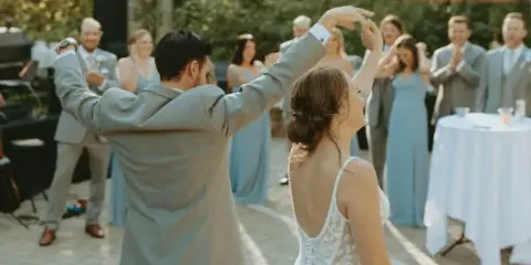 A bride and groom dancing at an outdoor wedding reception while guests in formal attire watch and applaud.