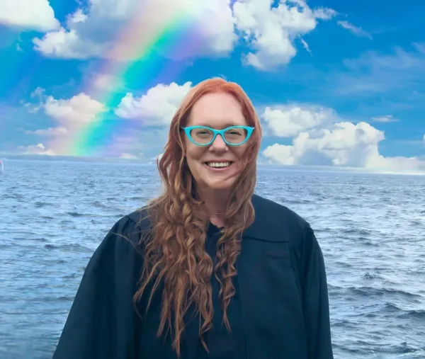 Portrait of a smiling woman with red hair and blue glasses standing in front of a body of water with a rainbow in the sky.