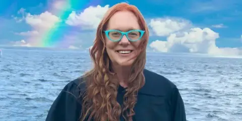 Portrait of a smiling woman with red hair and blue glasses standing in front of a body of water with a rainbow in the sky.