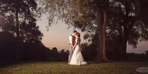A bride and groom standing together in a grassy field with large trees at sunset.