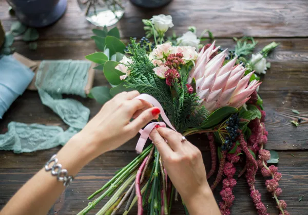 A close-up of a florist's hands tying a pink ribbon around a fresh floral bouquet containing protea and greenery on a wooden table.