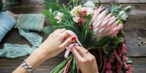 A close-up of a florist's hands tying a pink ribbon around a fresh floral bouquet containing protea and greenery on a wooden table.